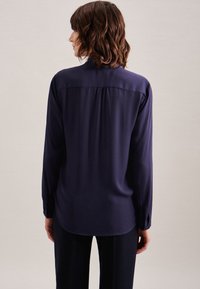 Blouse navy à manches longues avec une texture lisse, des poignets boutonnés et un design plissé au dos, associée à un pantalon noir ample.