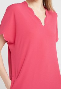 Rosa Bluse aus einem leichten, glatten Stoff mit einem gewellten Ausschnitt und kurzen Ärmeln. Verfügt über Seitentaschen und eine lockere Passform.