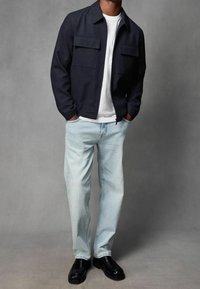 Giacca blu navy con zip e due tasche sul petto, abbinata a jeans a gamba dritta blu chiaro e mocassini neri. Tessuto liscio, design casual.