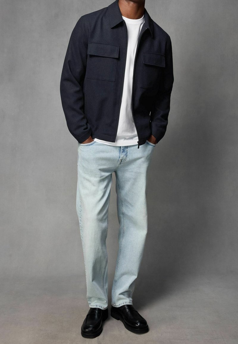 Giacca blu navy con zip e due tasche sul petto, abbinata a jeans a gamba dritta blu chiaro e mocassini neri. Tessuto liscio, design casual.