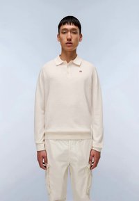 Svijetli krem pulover s dugim rukavima, ovratnikom i malim crvenim logotipom. Nošen uz odgovarajuće svijetle krem cargo hlače. Glatka tekstura.