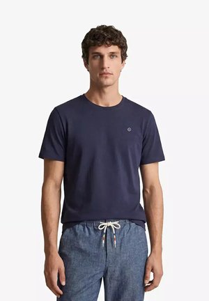 Uomo con capelli ricci che indossa una maglietta blu navy e pantaloni azzurri con coulisse, in piedi davanti a uno sfondo bianco semplice.