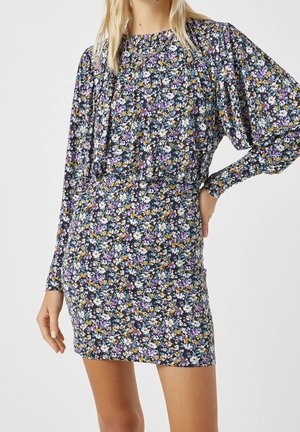 Femme portant une mini-robe ajustée à manches longues avec un motif floral bleu, blanc, violet et jaune sur un fond sombre.
