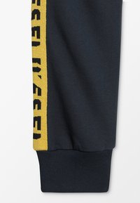 Marina sweatpants med en gul sidostrip som har "DIESEL" i svart text och elastiska muddar. Tillverkade av ett mjukt, texturerat material.