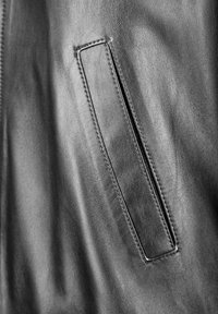 Close-up van een zwarte leren jas met een rechthoekige zak en stiksels. De textuur is glad met een licht glanzende afwerking.