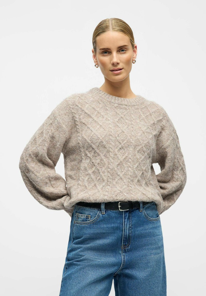 Suéter de punto aguja beige con cuello redondo y mangas largas y voluminosas, combinado con unos jeans de denim azul y un cinturón negro.