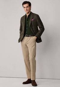 Blazer à carreaux marron porté sur une chemise vert foncé, chinos beiges et bottines marron. Comprend une pochette violette. Textures lisses.
