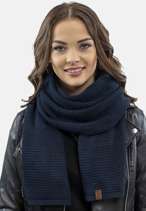 Écharpe en tricot bleu marine avec une surface texturée, enroulée autour du cou, présentant une forme rectangulaire et une petite étiquette de marque à l'extrémité.