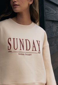 Beige crewneck sweatshirt met bordeauxrode geborduurde tekst "SUNDAY" en "Social Society since 1982" met geribbelde halslijn en manchetten.