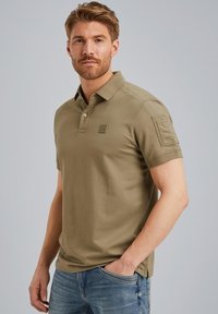 Kaki poloshirt met korte mouwen, drie knopenrij en een klein geborduurd logo op de borst. Draagt samen met lichtblauwe spijkerbroek.