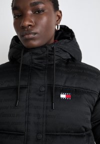 Black padded jacket with a textured fabric, featuring a logo patch and drawstrings on the hood. Buttons line the front for closure.