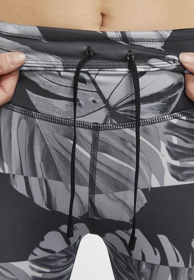 Ręce dostosowujące pas w szarych i czarnych legginsach w wzór, z sznurkiem do ściągania, odsłaniające brzuch i pępek.