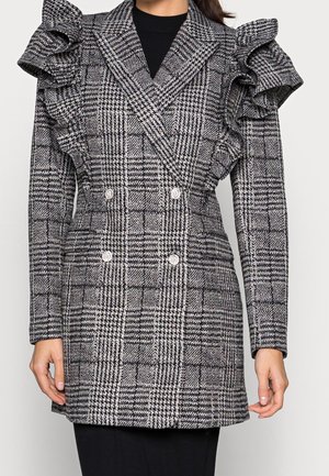 Veste croisée noir et blanc à carreaux avec épaulettes à volants portée sur un haut noir, montrée du cou jusqu'à la mi-cuisse.