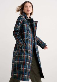 Cappotto trench a quadri blu scuro, con linee verdi, rosse e beige, due tasche frontali e bottoni grandi. Tessuto texturizzato con lucentezza visibile.