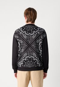 Sudadera negra con un contraste de patrones de telaraña blanca y bandana en la parte posterior. Presenta puños acanalados y cuello redondo.
