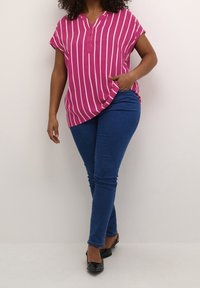 Blouse à manches courtes rayée rose et blanche avec un col en V et des boutons, associée à un jean skinny bleu et des chaussures noires.