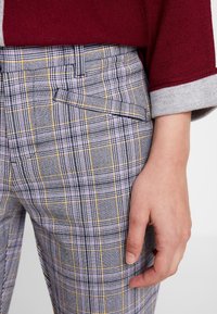 Main reposant sur un pantalon à carreaux jaunes et violets, portant un pull bordeaux et gris avec une manche retroussée.