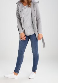 Jasnoszary płaszczyk z kapturem, warstwowy na szarym topie. Niebieskie wąskie dżinsy i białe trampki dopełniają stylizację. Widoczne minimalne wzory.