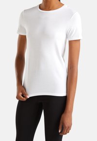 Kvinna klädd i en enkel vit kortärmad t-shirt med rund hals och svarta leggings, som håller i nederkanten av t-shirten med ena handen.