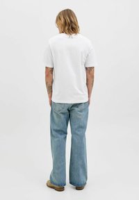 Biała bawełniana koszulka z krótkimi rękawami połączona z jasnoniebieskimi, szerokimi spodniami dżinsowymi, stojąca na tle jednolitego białego tła.