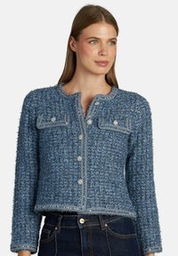 Giacca in tweed blu con due taschini sul petto, chiusura con bottoni metallici e design accorciato. Presenta una finitura texturizzata e un collo rotondo.