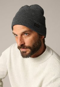 Bonnet gris tricoté avec texture côtelée et un patch logo en cuir noir. Porté par une personne portant un pull côtelé de couleur claire.