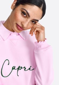 Felpa rosa chiaro con la scritta "Capri" ricamata in verde. Il colletto è bianco con un bottone e le maniche hanno polsini arricciati.