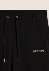 Pantalons de survêtement noirs avec une taille élastique, des cordons de serrage noirs et un logo "FREDDY" en argent métallisé. Texture lisse avec une coupe ajustée.