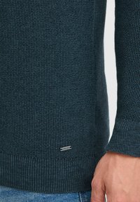 Knit sweater in dark blue with textured, ribbed cuffs and hem. Features a small silver tag reading "INDICODE" on the side.