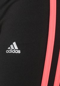 Czarnie spodnie sportowe z różowymi paskami po bokach, z małym białym logo Adidas na dolnej części nogawki. Gładka tekstura i dopasowany krój.