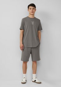 Junger Mann steht mit den Händen in den grauen Shorts-Taschen, trägt ein graues T-Shirt mit weißem Text und weiße Sneaker vor einem einfarbigen Hintergrund.
