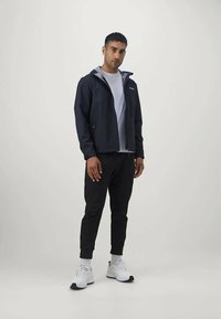 Mornarska vodoodporna jakna s sivo kapuco in zadrgo, v kombinaciji s črnimi ozkimi hlačami in belimi športnimi čevlji. Sproščen mestni outfit.