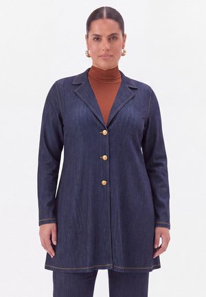 Kvinde iført en mørkeblå denim lang blazer med guldknapper over en brun rullekrave, stående mod en ensfarvet hvid baggrund.