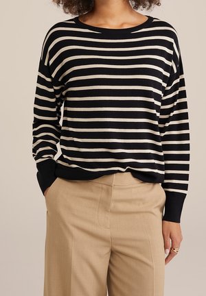 Nainen pukeutuneena mustavalkoraidalliseen pitkähihainen yläosaan, joka on työnnetty beigen värisiin korkeavyötäröisiin housuihin, käsi taskussa, seisoo yksiväristä taustaa vasten.