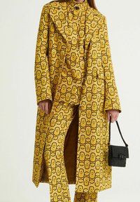 Trench giallo con motivo a disegni geometrici audaci, abbinato a pantaloni coordinati e a una borsa nera testurizzata.