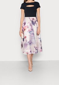 Haut noir avec une découpe à l'avant, associé à une jupe mi-longue ornée d'un motif floral dans des couleurs pastels. Les chaussures sont nude et il y a une petite pochette.