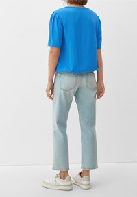 Blaues kurzärmliges Oberteil mit gerafftem Schulterdetail, kombiniert mit hellblauen geraden Jeans und weißen Turnschuhen. Einfaches, lässiges Outfit.