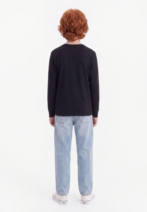 Barn med krøllet rødt hår står med ryggen til iført en sort langærmet skjorte, lyseblå jeans og hvide sneakers på en hvid baggrund.