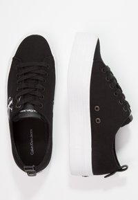 Baskets en toile noire avec une semelle en caoutchouc blanche, dotées de lacets ronds, d'œillets latéraux et d'une semelle intérieure de marque.