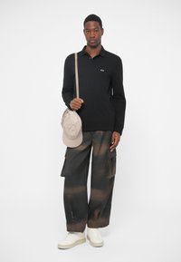 Homem a usar polo preto de mangas longas, calças cargo largas em preto e castanho, ténis brancos, segurando a alça de uma mala beige sobre o ombro, em fundo branco.