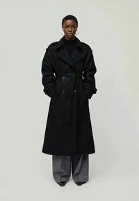 Cappotto trench nero con design avvolgente, frontale doppiopetto e cintura. Il modello indossa pantaloni grigi larghi e scarpe nere con finitura opaca.