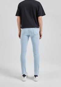 Jean skinny bleu clair à texture lisse, associé à une chemise noire à manches courtes et des baskets blanches. La vue arrière met en valeur les détails des poches.