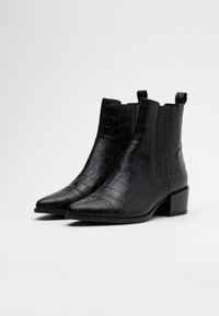 Botines negros de tobillo hechos de cuero sintético texturizado imitando cocodrilo, con punta afilada y tacón bajo, que cuentan con paneles laterales elásticos y lengüetas para facilitar su colocación.