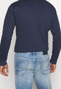 Man som bär en marinblå skjorta med lång ärm instoppad i ljusblåa urtvättade jeans, sedd bakifrån med avslappnade händer vid sidorna.