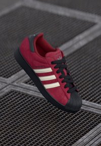 Red suede sneaker with black rubber sole, featuring three white stripes and black laces. Textured accents on tongue and toe cap.