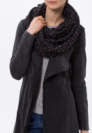 Femme portant une écharpe infinie noire à motifs triangulaires, un pull gris, un manteau gris foncé et un jean bleu, tenant l'écharpe près du cou.