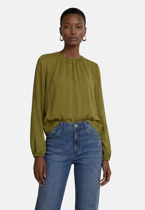 Femme portant un chemisier vert olive à manches longues et un jean bleu taille haute, debout devant un fond clair uni.