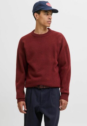 Maglione a maglia bordeaux con scollo rotondo e polsini a coste, abbinato a pantaloni blu scuro e un cappellino navy con una toppa con logo.