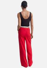 Zwarte tanktop en rode wijde broek. De katoenen broek heeft een elastische tailleband en een ontspannen pasvorm. Opmerkelijke tatoeage op de rug.