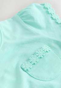 Krátky rukáv s mintovo zeleným tričkom s okrúhlym výstrihom, zdobený kvetinovou výšivkou na vrecku a šve na ramene. Hladká textúra látky.